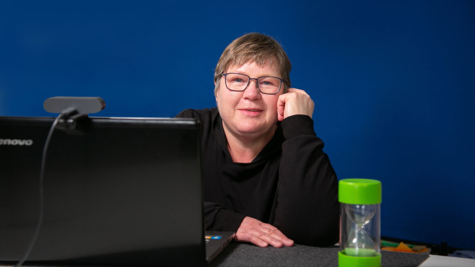 Jutta Wergen mit Laptop und Büchern auf dem Sofa Jutta Wergen sitzt an einem Laptop mit Webcam und stützt den Kopf auf die Hand. Neben ihr steht eine grüne Sanduhr.
